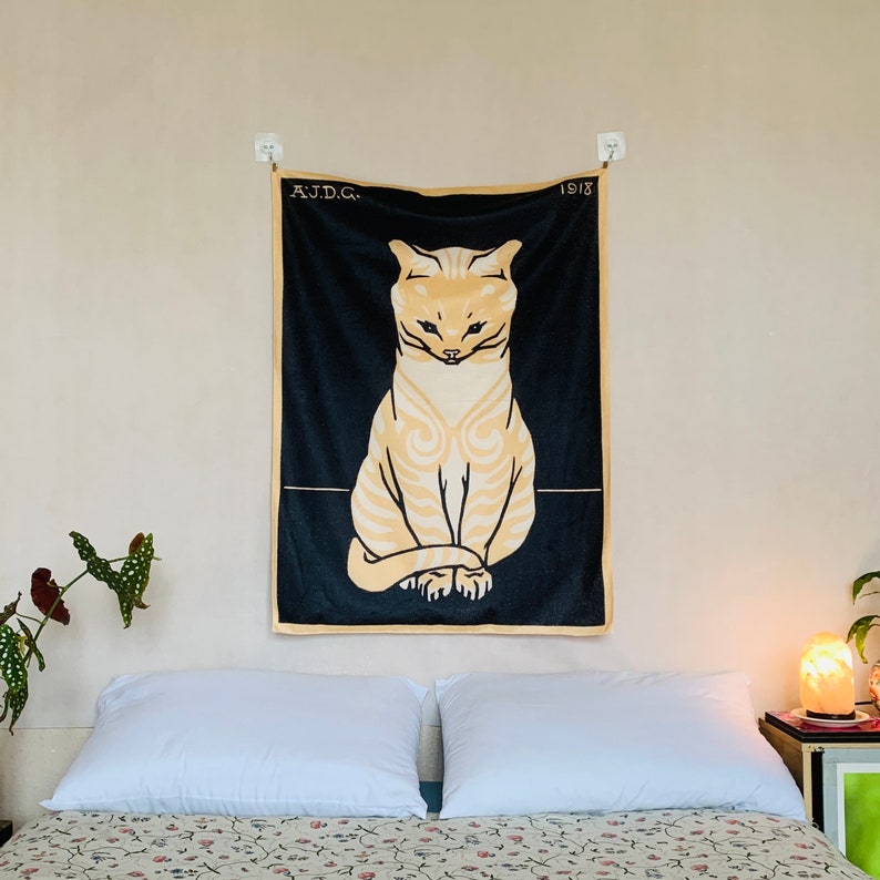 A Julie de Graag Sitting Cat tapestry hanging straight above a bed with vintage floral sheets. The black and yellow animal art wall hanging is styled next to a glowing salt lamp and a leafy green houseplant.