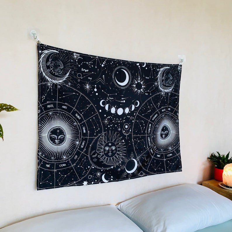 Black and white celestial zodiac tapestry hanging above a bed, featuring moon phases, constellations, and sun moon faces.