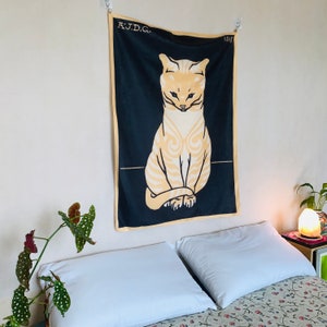 Angled view from the right of an animal art decor tapestry displayed in a cozy bedroom. The vintage 1918 illustration of a seated cat is softly illuminated by a Himalayan salt lamp resting on the bedside table.