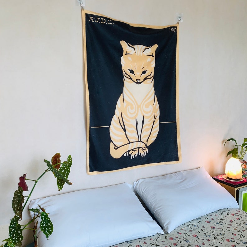 Angled view from the right of an animal art decor tapestry displayed in a cozy bedroom. The vintage 1918 illustration of a seated cat is softly illuminated by a Himalayan salt lamp resting on the bedside table.