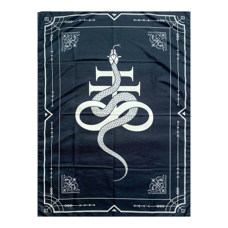 Op de afbeelding: Een zwart wandkleed met een witte slang die om een kruis is gewikkeld. Het wandkleed heeft een decoratieve rand.