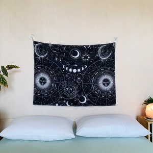 Wide room view of black and white zodiac tapestry above a bed, featuring moon phases, constellations, and celestial sun moon design.
