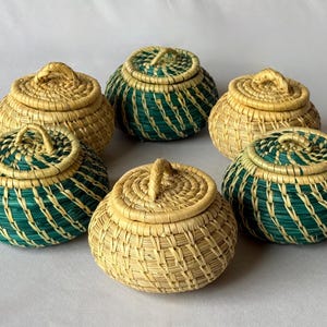May include: A collection of six small, round, lidded baskets arranged in a circle. Three baskets are natural light tan, and three are a combination of natural tan and dark teal green. Each basket features a woven lid with a small central handle.