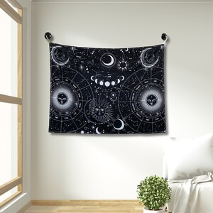 Black and white celestial astrology tapestry hanging on a wall, with moon phases, zodiac circles, constellations, and sun moon faces.