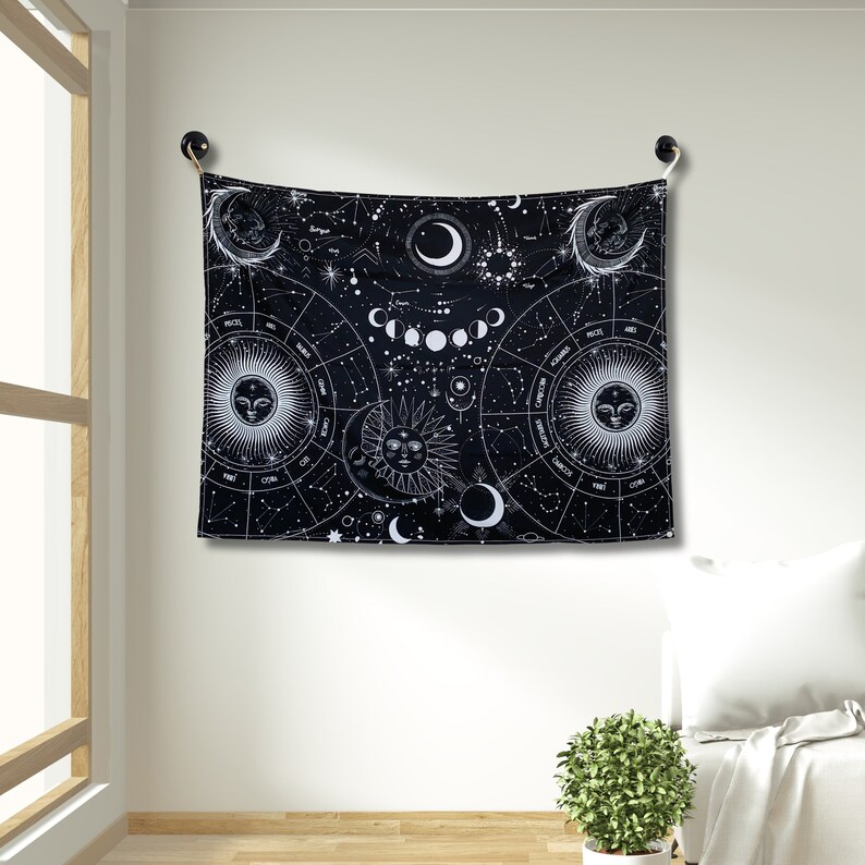 Black and white celestial astrology tapestry hanging on a wall, with moon phases, zodiac circles, constellations, and sun moon faces.
