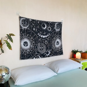 Angled bedroom view of black and white celestial zodiac tapestry, featuring moon phases, constellation patterns, and mystical sun moon imagery.