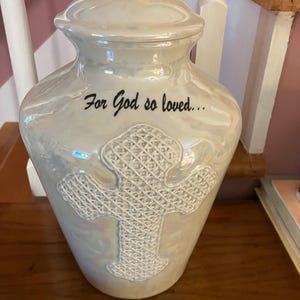 May include: A pearlescent white ceramic urn with a removable lid, featuring a raised textured cross design on the front. The text "For God so loved..." is printed in black script above the cross. The surface has a glossy, iridescent finish.