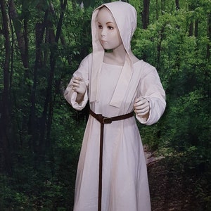 May include: A white linen dress with a long, flowing skirt and a fitted bodice. The dress has a hood and a brown leather belt. The dress is worn by a mannequin in a forest setting.
