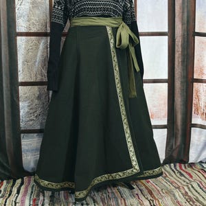 May include: A black sweater with a geometric pattern and a dark green wrap skirt. The skirt has a decorative trim and a matching green sash tied at the waist. The outfit is displayed in front of a window.