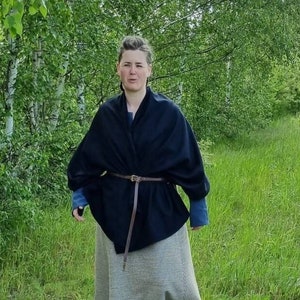 May include: A woman wearing a black shawl with a brown leather belt over a gray skirt, standing in a grassy area with trees in the background.