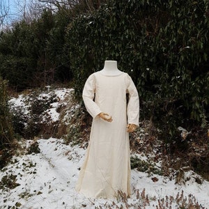 May include: A long, white linen dress with a round neckline and long sleeves. The dress is displayed on a mannequin in a snowy outdoor setting.