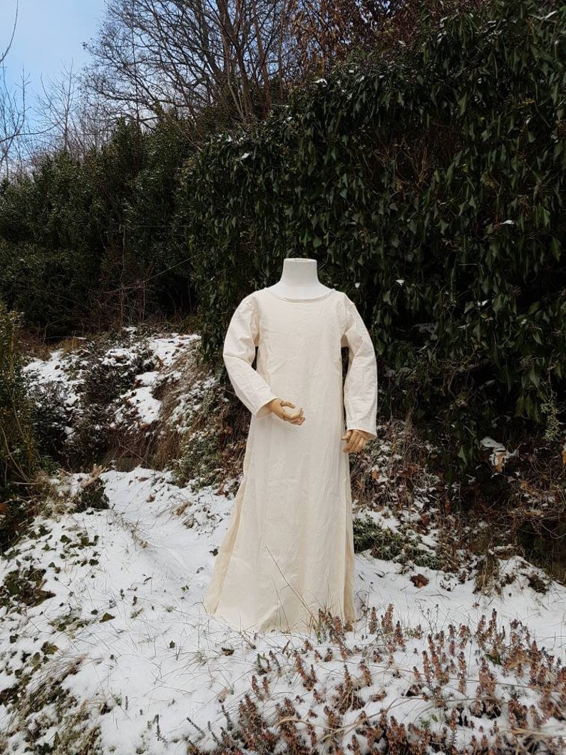 May include: A long, white linen dress with a round neckline and long sleeves. The dress is displayed on a mannequin in a snowy outdoor setting.