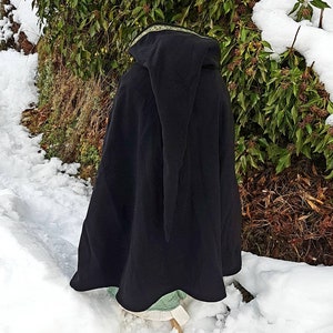 May include: Black hooded cloak with a gold trim, worn over a green and white dress. The person is standing in a snowy landscape.