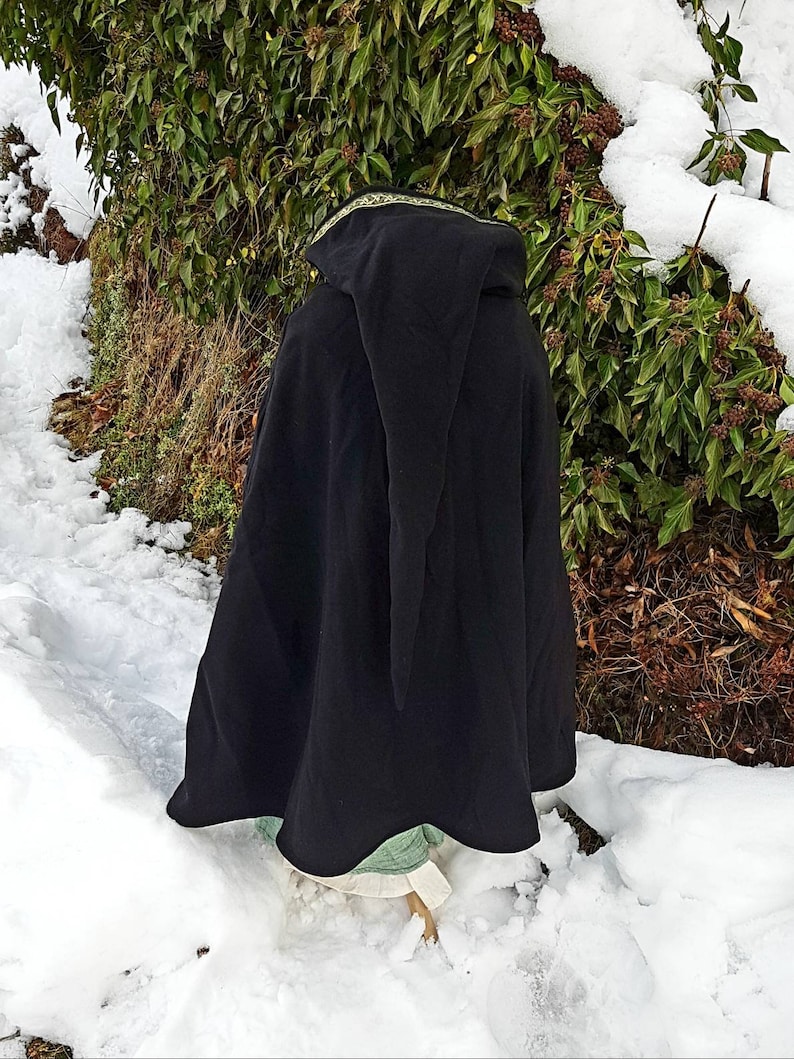 May include: Black hooded cloak with a gold trim, worn over a green and white dress. The person is standing in a snowy landscape.