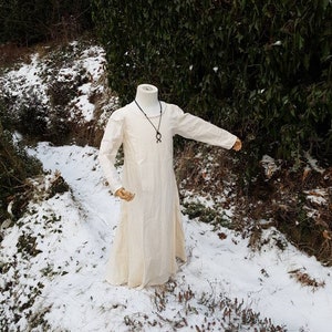 May include: A white linen tunic with long sleeves, worn on a mannequin. The tunic has a simple, loose fit and is shown against a snowy background.