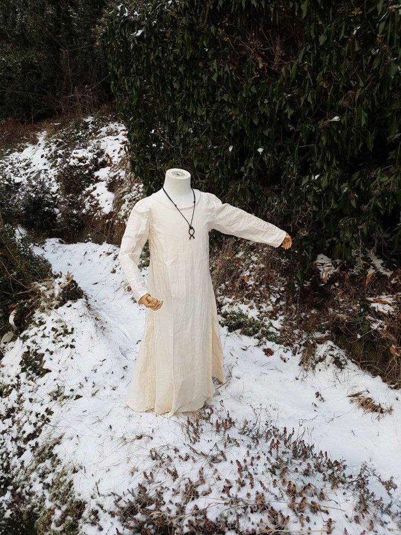 May include: A white linen tunic with long sleeves, worn on a mannequin. The tunic has a simple, loose fit and is shown against a snowy background.