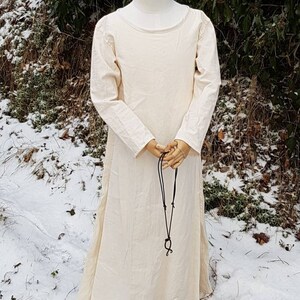 May include: A long, white linen dress with long sleeves and a round neckline. The dress is worn on a mannequin and has a black leather cord hanging from the neckline.