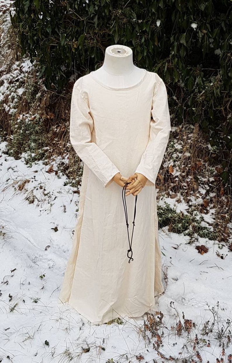 May include: A long, white linen dress with long sleeves and a round neckline. The dress is worn on a mannequin and has a black leather cord hanging from the neckline.