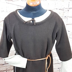 May include: A brown tunic with long sleeves and a round neckline. The tunic has a brown leather belt with a long strap hanging down the front. The sleeves are gathered at the wrists and have a white underlayer.