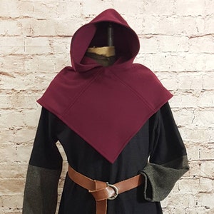 Könnte beinhalten: Ein burgunderfarbener Kapuzenumhang mit einer spitzen Kapuze und einer schwarzen Tunika darunter. Der Umhang ist aus einem weichen, warmen Stoff gefertigt und hat ein schlichtes, elegantes Design. Die Tunika hat einen braunen Ledergürtel mit einer silbernen Schnalle.