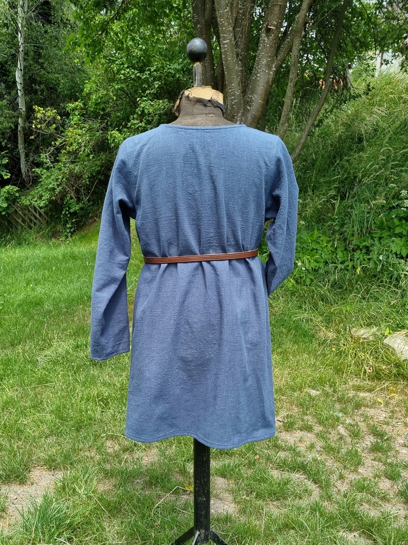 May include: A long-sleeved, blue tunic with a brown belt. The tunic is made of a textured fabric and has a simple, classic design.