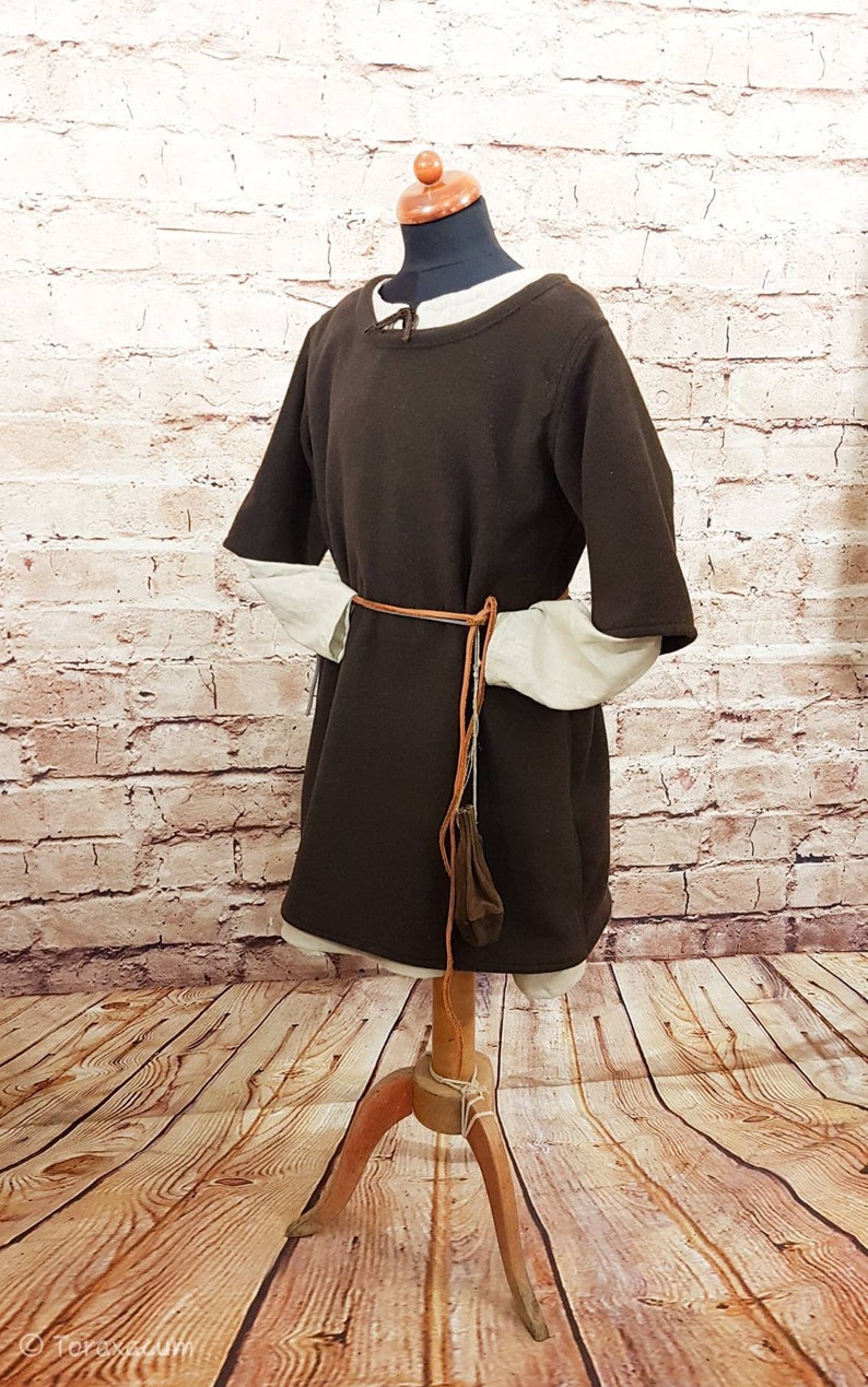 May include: A brown tunic with short sleeves and a brown leather belt. The tunic is on a wooden mannequin against a white brick wall.