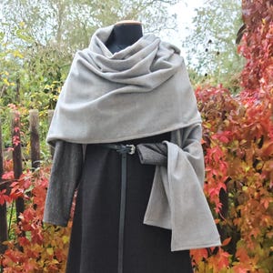 Op de afbeelding: Een grijze wollen cape en sjaal set gepresenteerd op een mannequin. De cape is donkergrijs en de sjaal is lichtgrijs. Een zwarte riem sluit de cape in de taille. De achtergrond toont een houten hek en kleurrijk herfstblad.