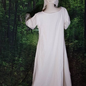 May include: A white linen dress with long sleeves and a gathered neckline. The dress is worn by a mannequin in a forest setting.