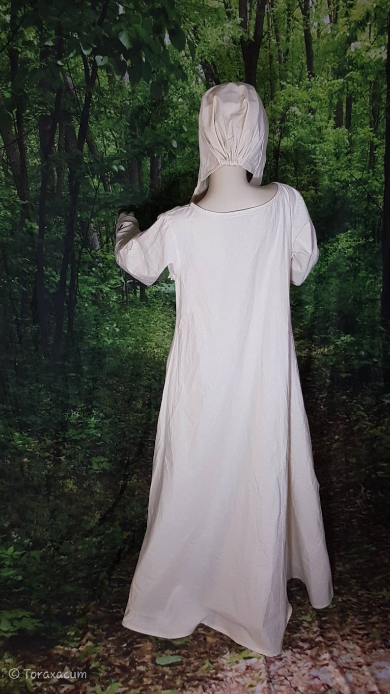 May include: A white linen dress with long sleeves and a gathered neckline. The dress is worn by a mannequin in a forest setting.