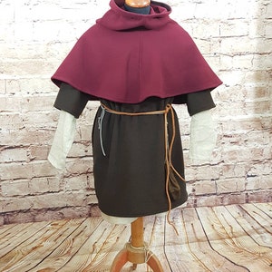 May include: A maroon hooded cloak with a brown tunic underneath. The cloak has a wide, rounded collar and is worn over a brown tunic with a leather belt. The mannequin is standing on a wooden floor.