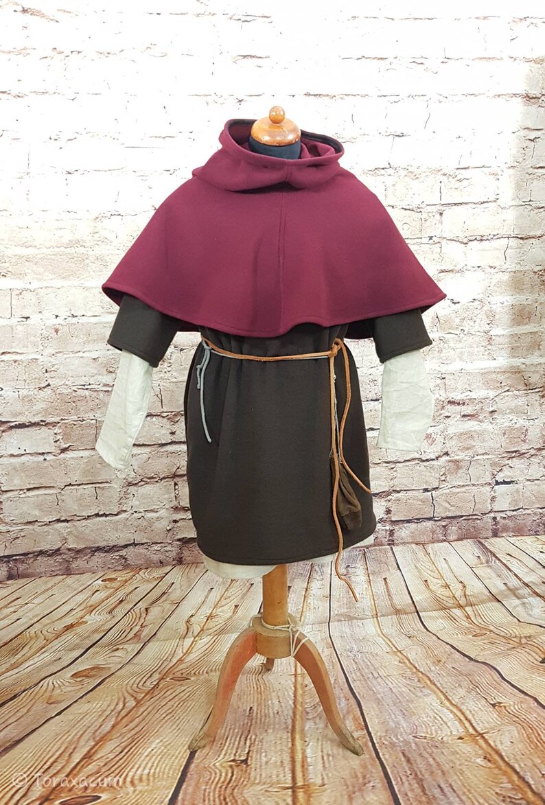 May include: A maroon hooded cloak with a brown tunic underneath. The cloak has a wide, rounded collar and is worn over a brown tunic with a leather belt. The mannequin is standing on a wooden floor.