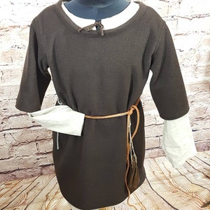 May include: A brown tunic with long sleeves and a round neckline. The tunic is belted with a brown leather belt and has a white linen undergarment peeking out from the sleeves.