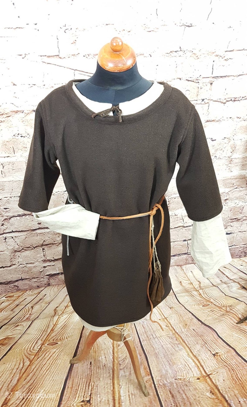 May include: A brown tunic with long sleeves and a round neckline. The tunic is belted with a brown leather belt and has a white linen undergarment peeking out from the sleeves.