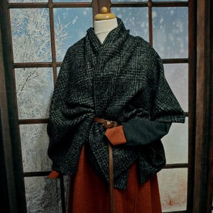 May include: A dark gray and black plaid cape with a brown leather belt, displayed on a mannequin. The cape is draped over a rust-colored skirt and dark green sleeves. The background shows a window with a winter scene.