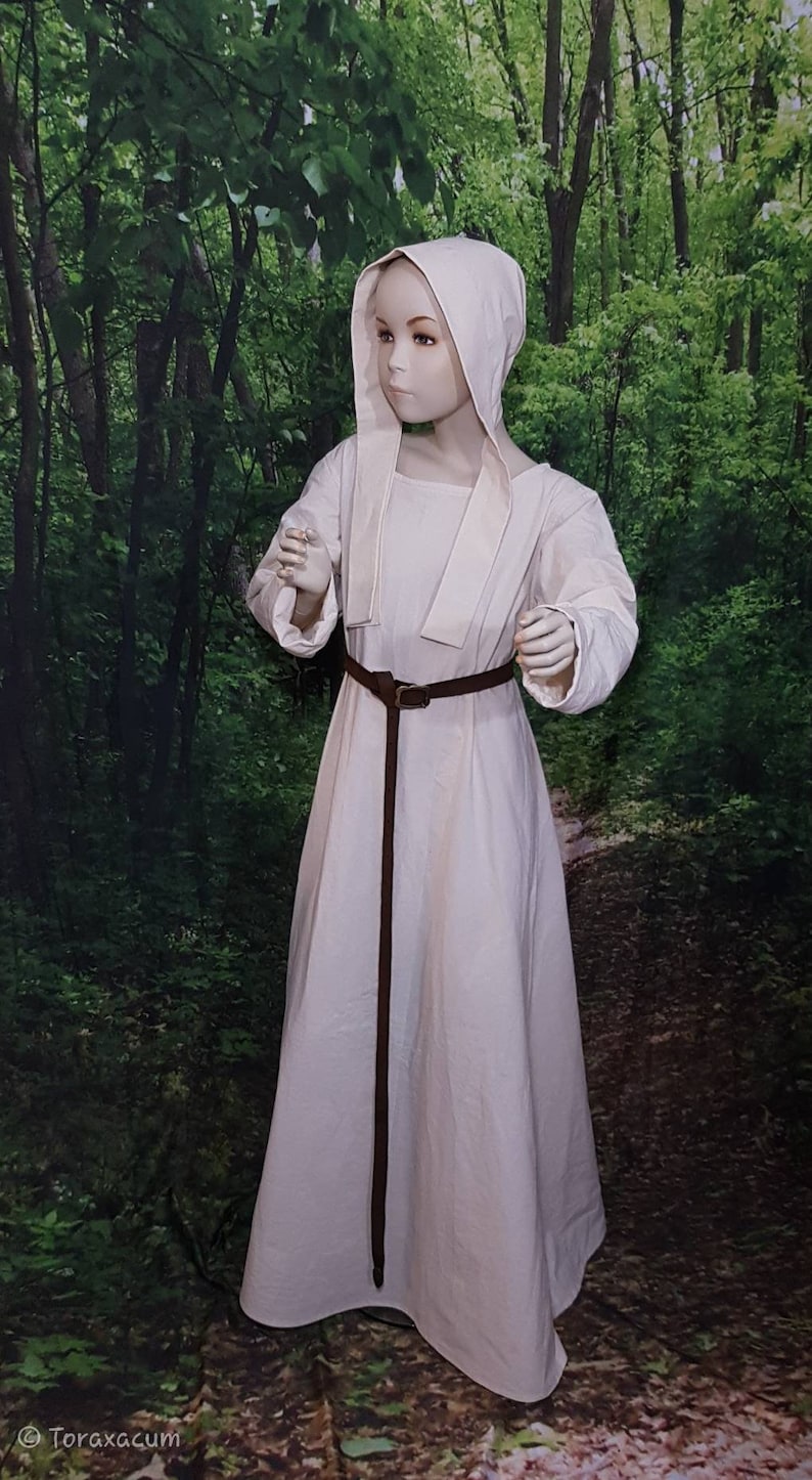 May include: A white linen dress with a long, flowing skirt and a fitted bodice. The dress has a hood and a brown leather belt. The dress is worn by a mannequin in a forest setting.