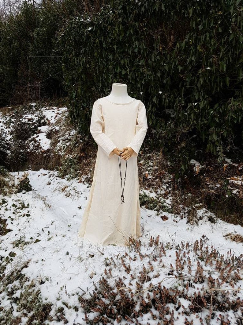 May include: A white linen tunic with long sleeves and a round neckline. The tunic is worn on a mannequin and is standing in a snowy landscape.