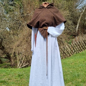 Op de afbeelding: Een bruine capuchon met een leren riem en een witte linnen jurk. De capuchon wordt over de jurk gedragen en de riem is in de taille geknoopt.
