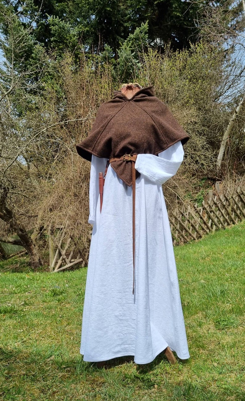 Op de afbeelding: Een bruine capuchon met een leren riem en een witte linnen jurk. De capuchon wordt over de jurk gedragen en de riem is in de taille geknoopt.