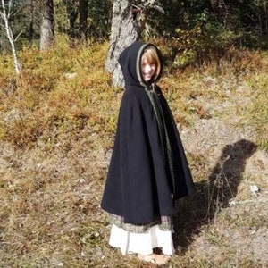 May include: A black hooded cloak with a green trim. The cloak is worn by a person standing in a wooded area.