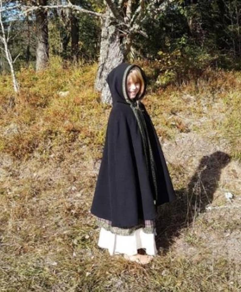 May include: A black hooded cloak with a green trim. The cloak is worn by a person standing in a wooded area.