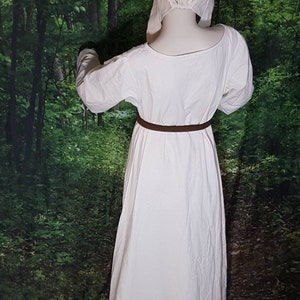 May include: A white linen dress with long sleeves and a brown belt. The dress is worn by a person with a white linen cap on their head. The person is standing in a forest setting.