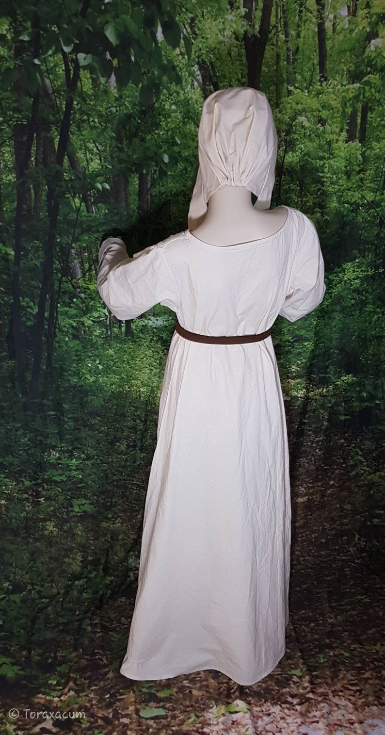 May include: A white linen dress with long sleeves and a brown belt. The dress is worn by a person with a white linen cap on their head. The person is standing in a forest setting.