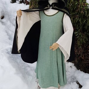 May include: A black hooded cloak with a green and gold trim, worn over a green linen dress with white sleeves. The dress has a simple, A-line silhouette. The cloak and dress are both made of natural fabrics.