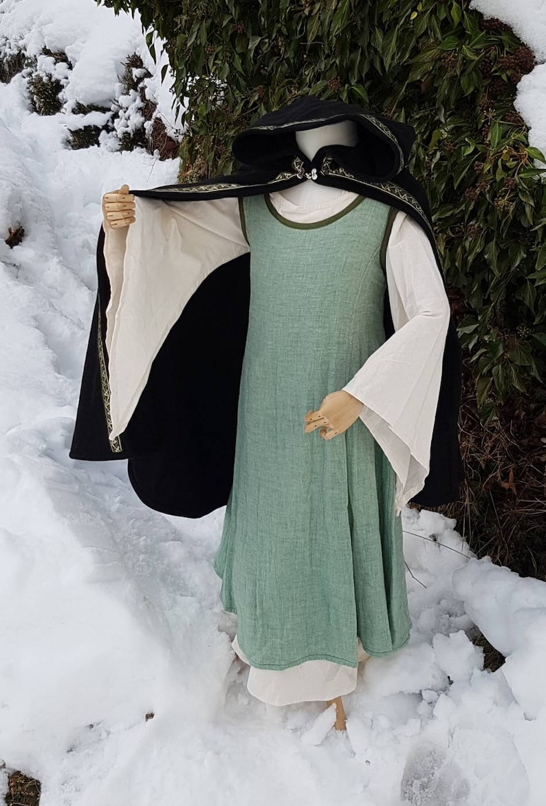 May include: A black hooded cloak with a green and gold trim, worn over a green linen dress with white sleeves. The dress has a simple, A-line silhouette. The cloak and dress are both made of natural fabrics.