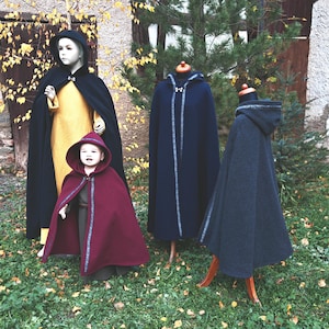 May include: A display of hooded capes in various colors. A black cape is worn by a mannequin in a yellow dress. A child wears a burgundy cape with a matching hood. Two additional capes are displayed on mannequins, one navy blue and one gray.