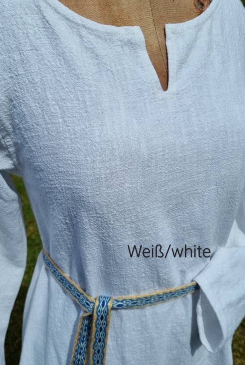 Op de afbeelding: Een witte linnen tuniek met een V-hals en lange mouwen. De tuniek is in de taille gebonden met een geweven riem in blauw en beige. De tekst "Wei&szlig;/white" is zichtbaar op de stof.