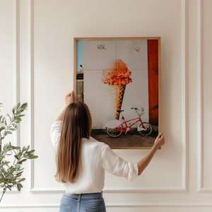 May include: A framed print featuring a large ice cream cone and a red bicycle. The artwork has a light brown wooden frame and is being held against a white wall. The ice cream is orange and the bicycle has white tires.