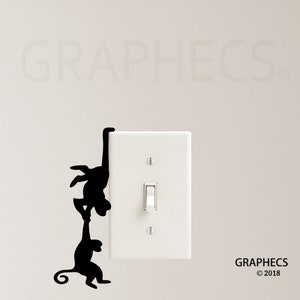 May include: Black monkey silhouette wall decal climbing on a white light switch cover. The decal features two monkeys, one hanging from the top and the other holding onto the first monkey's hand. The word "GRAPHECS" is in the background.