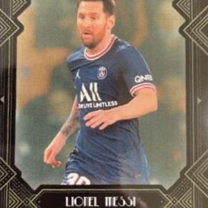 May include: A trading card featuring Lionel Messi in a dark blue soccer jersey with red and white trim. The card has a decorative black border with the text "Lionel Messi" and the number "08/99". The card also has the Topps logo.