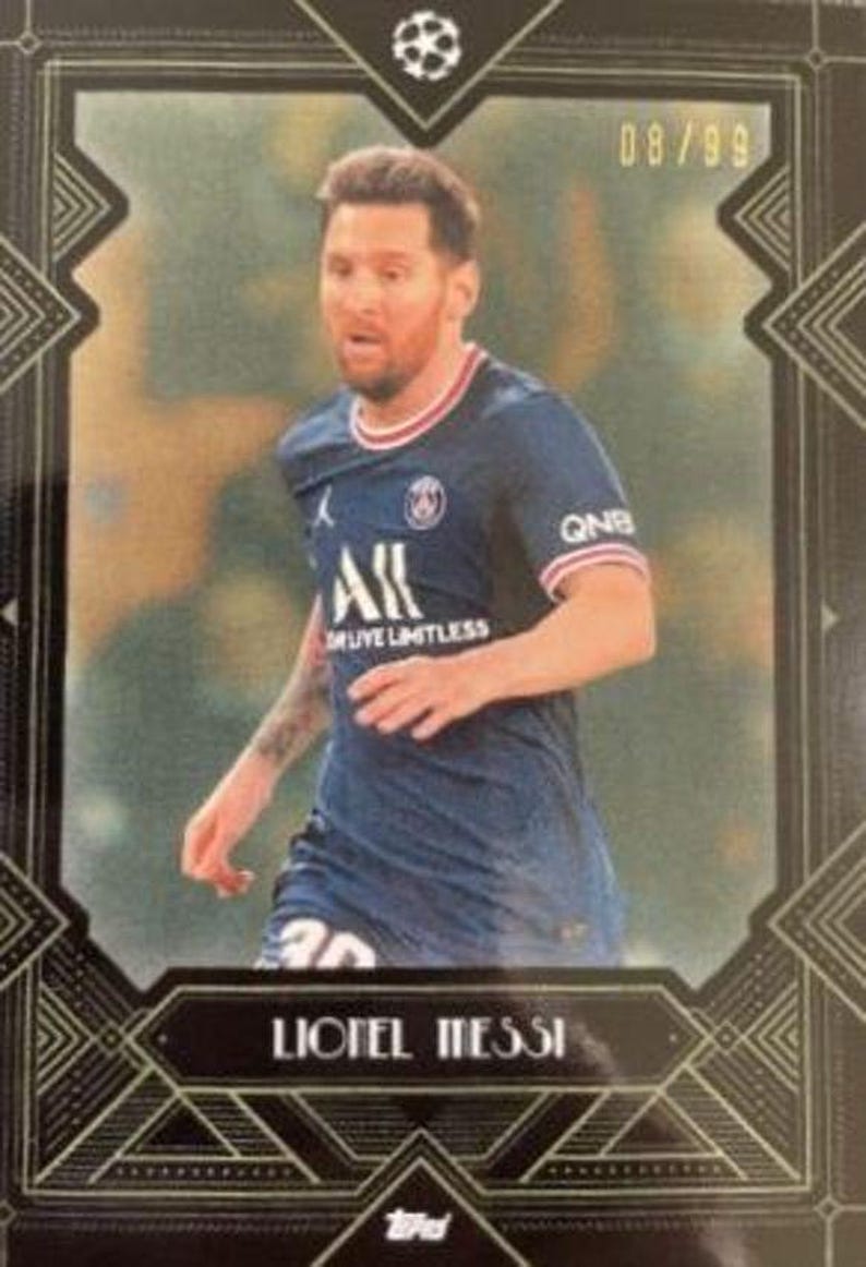 May include: A trading card featuring Lionel Messi in a dark blue soccer jersey with red and white trim. The card has a decorative black border with the text "Lionel Messi" and the number "08/99". The card also has the Topps logo.